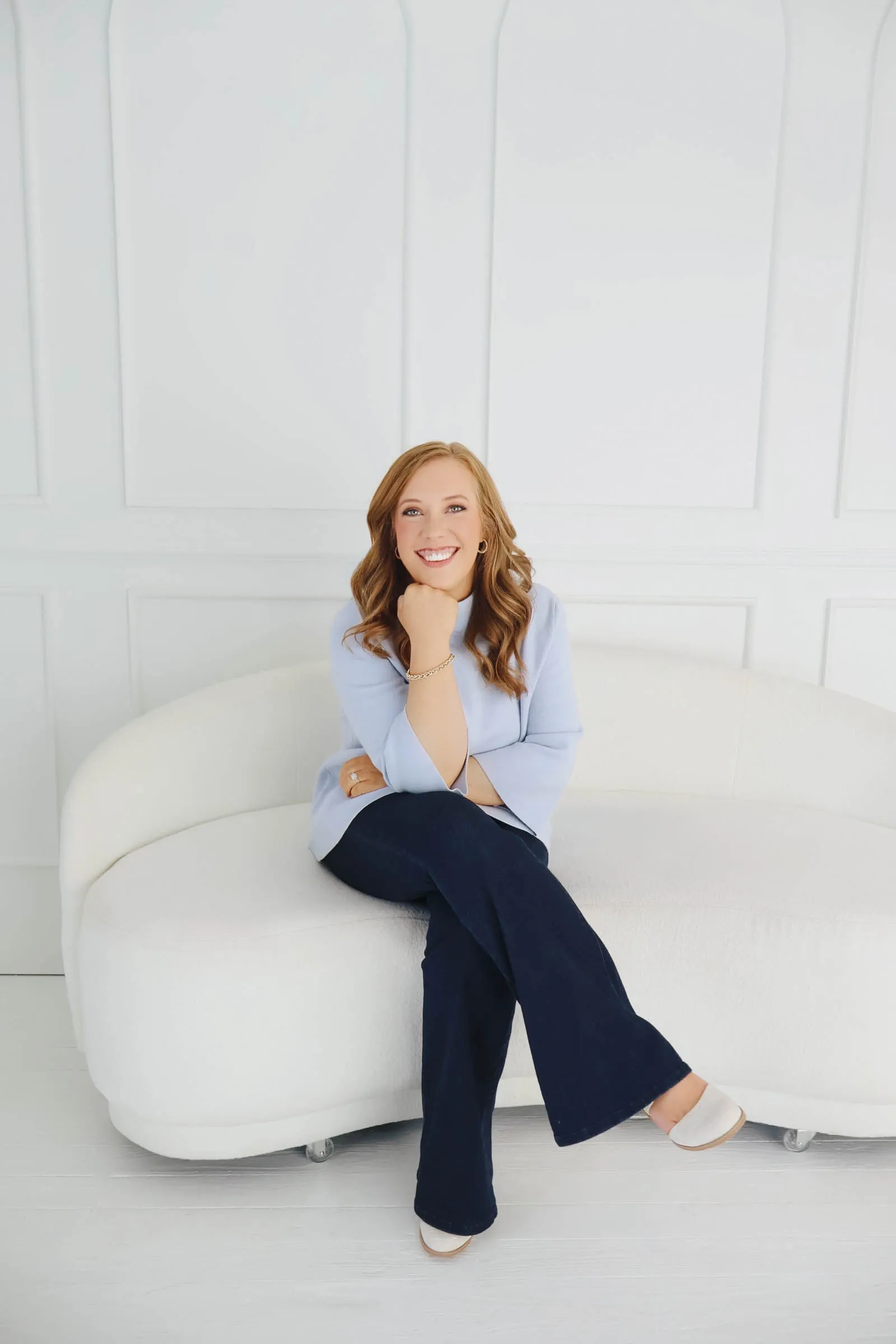 Kendra Madewell smiling on a white curved couch in a light blue sweater