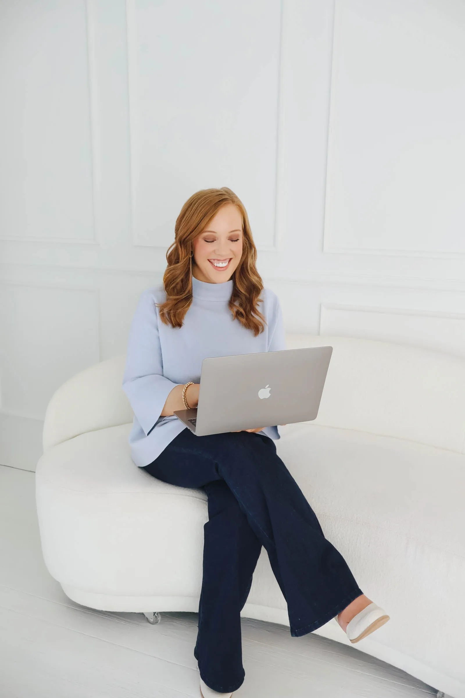 Kendra Madewell, a Northwest Arkansas buyer’s agent, smiling down at her laptop on a curved white couch